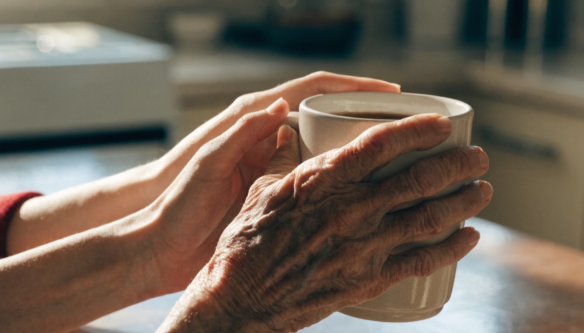A caregiver steadying an elderly person's hands around a warm mug